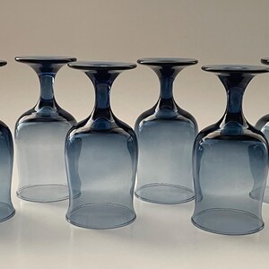 May include: Six blue-tinted wine glasses are arranged in a row, each with a rounded bowl and a flared rim. The stems and rims are a darker shade of blue, creating a gradient effect. The glasses are made of clear glass.