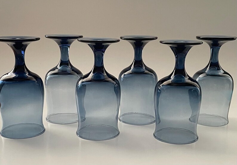 May include: Six blue-tinted wine glasses are arranged in a row, each with a rounded bowl and a flared rim. The stems and rims are a darker shade of blue, creating a gradient effect. The glasses are made of clear glass.