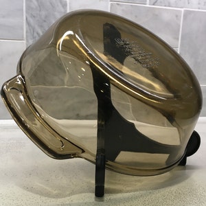 May include: A transparent, amber-colored glass baking dish with a rounded rectangular shape. The dish has two handles and is resting on a black stand. The dish appears to be empty and clean.