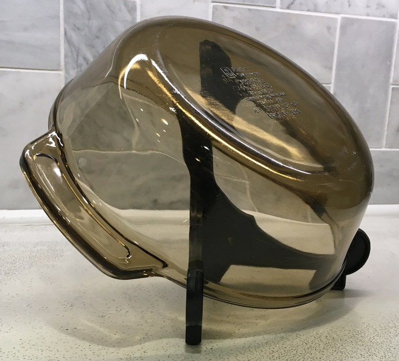 May include: A transparent, amber-colored glass baking dish with a rounded rectangular shape. The dish has two handles and is resting on a black stand. The dish appears to be empty and clean.