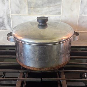 May include: A stainless steel cooking pot with a lid and two side handles. The pot is sitting on a black stovetop. The lid has a black handle. The bottom of the pot shows signs of wear.