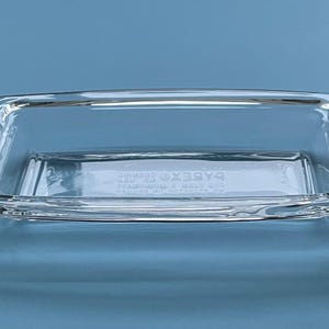 May include: A clear, rectangular glass butter dish with a handle. The dish has a slightly curved design and is made of transparent glass. The word "PYREX" is visible on the bottom of the dish. The dish is on a blue background.