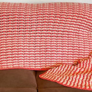 May include: A rectangular crochet blanket featuring a repeating textured pattern of alternating horizontal stripes in coral pink and cream yarn. The blanket has a finished edge in solid coral pink and displays a consistent, dense stitch design.