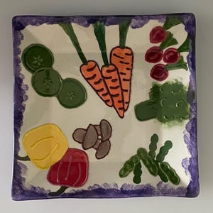 May include: Square ceramic plate with a white background and a purple border. The plate is decorated with colorful, hand-painted vegetables, including carrots, broccoli, and peppers. The design is whimsical and suitable for serving food.