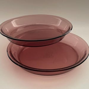 May include: Two transparent, oval-shaped, purple glass pie dishes are stacked on top of each other. The dishes have a slightly raised rim and a smooth surface. The color is a deep, rich purple.