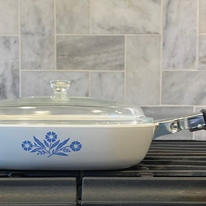 May include: A light blue casserole dish with a clear glass lid and a black handle. The dish features a blue floral design on the side. It is sitting on a black stove grate.
