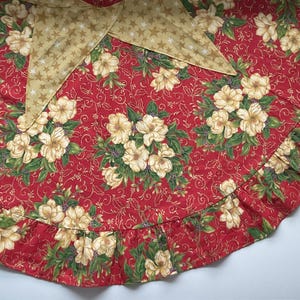 May include: A red Christmas tree skirt with a floral pattern and a gold star-patterned bow. The skirt features a ruffled edge and is decorated with cream-colored flowers and green leaves.