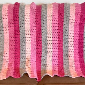 May include: A rectangular crochet blanket featuring vertical stripes in alternating shades of hot pink, light pink, and heather gray. The textile has a textured, repeating stitch pattern throughout and a scalloped edge along the top and bottom borders.