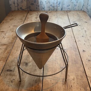 May include: A metal food mill featuring a conical perforated strainer supported by a wire stand. The device includes a wooden pestle with a rounded handle positioned inside the bowl, designed for pressing ingredients through the fine mesh holes.
