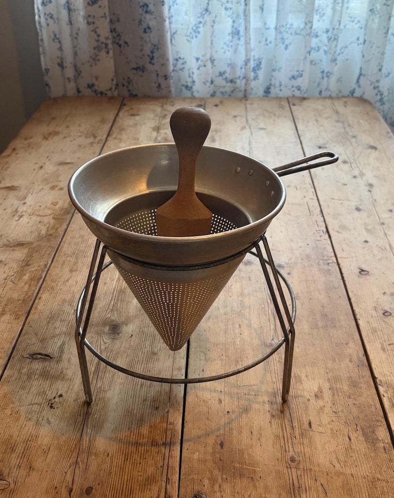 May include: A metal food mill featuring a conical perforated strainer supported by a wire stand. The device includes a wooden pestle with a rounded handle positioned inside the bowl, designed for pressing ingredients through the fine mesh holes.