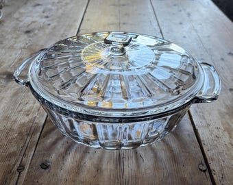 May include: A clear glass casserole dish with a matching lid, featuring a radial ribbed pattern on the lid and vertical fluted sides. The round dish includes two integrated side handles and a central square knob on the lid for easy lifting.
