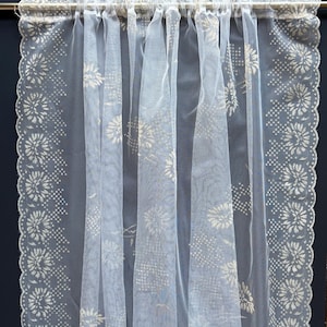 May include: Sheer white curtain with a floral lace border and a delicate, airy design. The curtain is hanging from a rod and features a repeating pattern of flowers and dots along the edges. The fabric is translucent, allowing light to filter through.