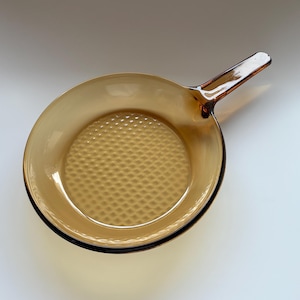 May include: Amber-colored glass frying pan with a long handle. The interior of the pan has a textured, diamond-patterned surface. The pan is designed for cooking and is made of durable glass.