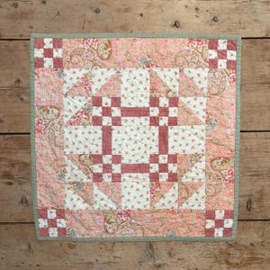 May include: A square quilted wall hanging featuring a central block pattern with dusty rose and cream floral fabrics. The design includes a patchwork border with small checkered corner squares and a sage green binding edge finish.