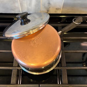 May include: A stainless steel saucepan with a copper-clad bottom and a matching lid. The bottom features an embossed Revere Ware logo with the text 1 QT, 96-A, Clinton, ILL, U.S.A. The pan has a long black handle with a metal hanging loop.