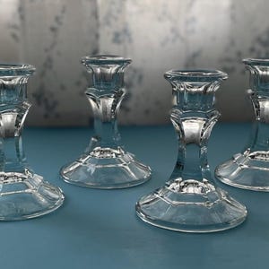 May include: Four clear glass candle holders with a classic design. Each holder has a flared base, a decorative stem, and a cup for holding a candle. The candle holders are arranged on a blue surface.