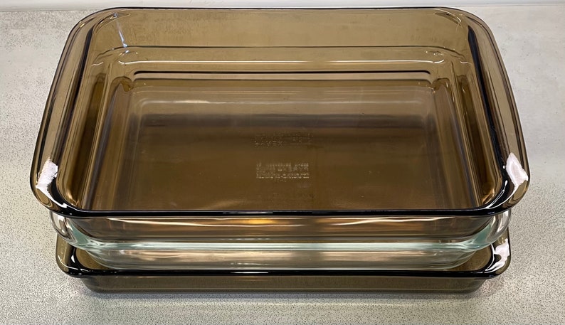 May include: Three rectangular, clear glass baking dishes stacked on top of each other. The dishes have a slightly tinted brown color. The bottom of the middle dish has the text "PYREX" and "MADE IN USA" imprinted.