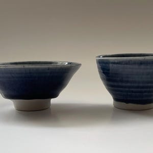 May include: Two blue ceramic bowls with a white base are displayed. The bowls have a textured, glazed finish. One bowl has a spout, and the other is round. The bowls are likely for serving food.