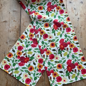 May include: A floral table runner featuring a white background with a repeating pattern of red roses, yellow sunflowers, and green leaves. The runner is made of fabric and is displayed on a wooden surface.