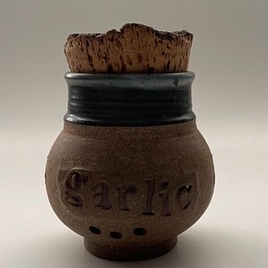 May include: A brown ceramic garlic keeper with a cork stopper. The jar has a rounded shape with the word "Garlic" carved into the side. It features a black band around the neck and small ventilation holes.