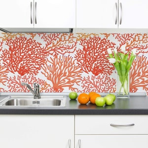 May include: A white kitchen with a backsplash featuring a coral reef design. The backsplash is made of white tiles with a red coral pattern. There are white cabinets above the backsplash and a stainless steel sink below. There are also some oranges and apples on the counter.