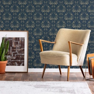 May include: A beige armchair with wooden legs sits in front of a wall covered in a dark blue wallpaper with a gold damask pattern. A framed picture and a potted plant are on the left side of the image.