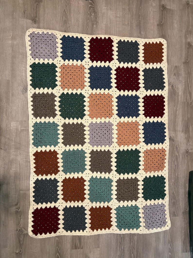 K&ouml;nnte beinhalten: Eine handgefertigte H&auml;keldecke mit Patchwork-Design. Die Decke besteht aus zahlreichen quadratischen Bl&ouml;cken in verschiedenen Farben, darunter Blau, Gr&uuml;n, Braun und Lila, mit einem cremefarbenen Rand. Die Decke liegt auf einer Holzoberfl&auml;che.