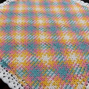 Puede incluir: Una manta cuadrada de ganchillo con un patrón variado de hilo amarillo, rosa y verde azulado intercalado con blanco. La manta tiene un punto texturizado en toda la superficie y está rematada con un borde decorativo de ganchillo blanco.