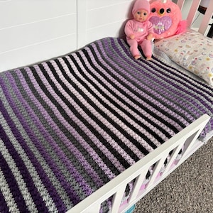 May include: A handmade crochet blanket in a crib. The blanket features alternating stripes of purple, gray, black, and white. A doll in pink pajamas and a pink unicorn pillow are on the crib.