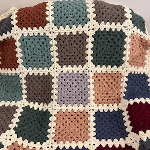 K&ouml;nnte beinhalten: Eine handgefertigte H&auml;keldecke mit Patchwork-Design. Die Decke zeigt Quadrate in verschiedenen Farben, darunter Braun, T&uuml;rkis und Burgunder, die von cremefarbenem Garn umrandet sind. Die Decke ist &uuml;ber eine Oberfl&auml;che drapiert.