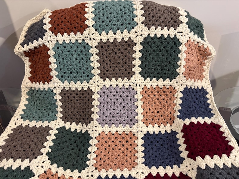 K&ouml;nnte beinhalten: Eine handgefertigte H&auml;keldecke mit Patchwork-Design. Die Decke zeigt Quadrate in verschiedenen Farben, darunter Braun, T&uuml;rkis und Burgunder, die von cremefarbenem Garn umrandet sind. Die Decke ist &uuml;ber eine Oberfl&auml;che drapiert.