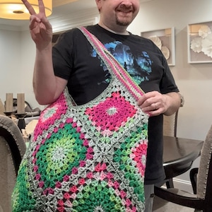 May include: A large, handmade crochet tote bag. The bag features a colorful granny square design with pink, green, and gray squares. The strap is a matching pink and gray. The bag is held by a person.