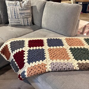K&ouml;nnte beinhalten: Eine handgefertigte H&auml;keldecke mit Patchwork-Design. Die Decke hat Quadrate in verschiedenen Farben, darunter Braun, Burgunderrot, Blau und Gr&uuml;n, mit einem cremefarbenen Rand. Sie liegt auf einem grauen Ecksofa.