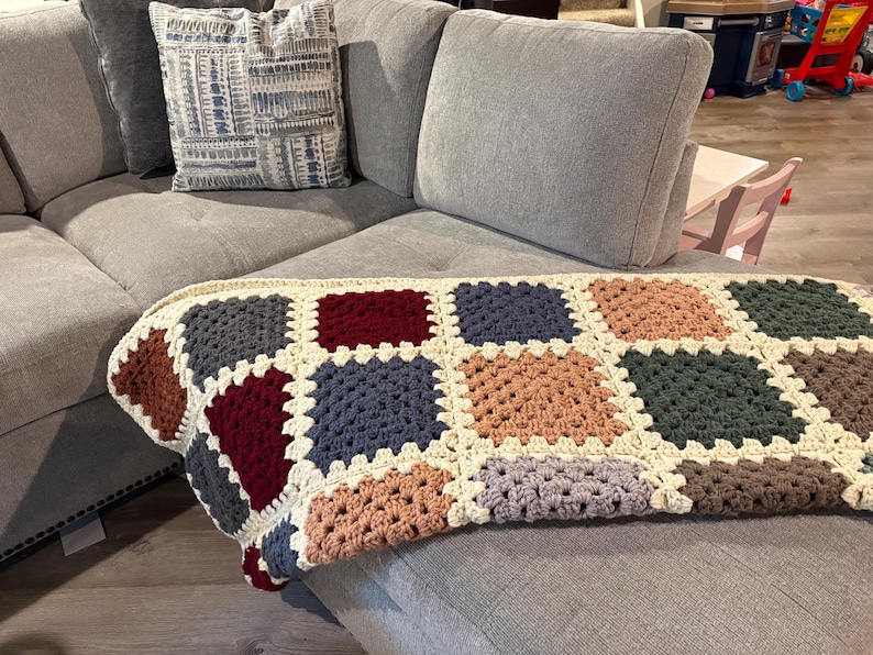 K&ouml;nnte beinhalten: Eine handgefertigte H&auml;keldecke mit Patchwork-Design. Die Decke hat Quadrate in verschiedenen Farben, darunter Braun, Burgunderrot, Blau und Gr&uuml;n, mit einem cremefarbenen Rand. Sie liegt auf einem grauen Ecksofa.