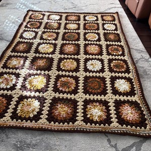May include: A rectangular crochet blanket featuring a grid of square motifs. Each square contains a circular floral pattern in shades of brown, cream, tan, and yellow, joined together by a cream-colored yarn border throughout the piece.