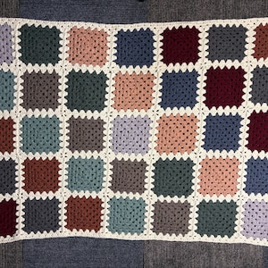 May include: A colourful, handmade crochet blanket with a patchwork design. The blanket is made up of squares in various colours, including blues, greens, browns, and pinks, with a white border. Ideal for adding a touch of warmth to any home.