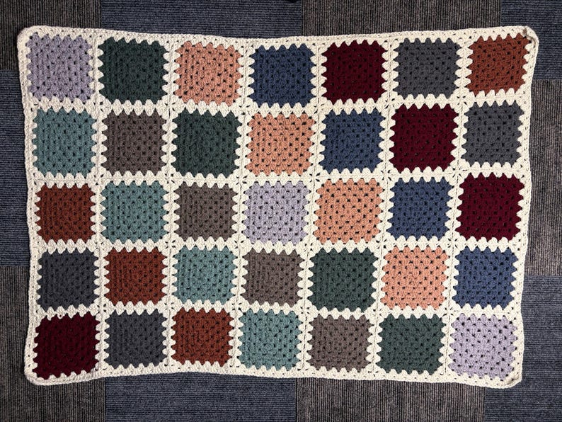 K&ouml;nnte beinhalten: Eine handgefertigte H&auml;keldecke mit Patchwork-Design. Die Decke besteht aus zahlreichen Quadraten in verschiedenen Farben, darunter Blau-, Gr&uuml;n-, Braun- und Rosat&ouml;ne, mit einem wei&szlig;en Rand. Ideal f&uuml;r die Inneneinrichtung.