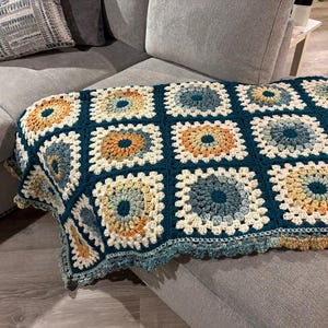 May include: A handmade crochet blanket in a granny square pattern. The blanket features squares in shades of teal, cream, orange, and yellow. The blanket is draped over a gray couch, showcasing its texture and design.