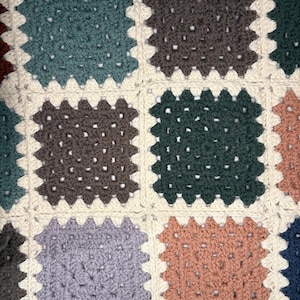 K&ouml;nnte beinhalten: Nahaufnahme einer handgefertigten H&auml;keldecke mit Patchwork-Design. Die Decke zeigt Quadrate in verschiedenen Farben, darunter T&uuml;rkis, Braun, Gr&uuml;n und Pfirsich, vor einem cremefarbenen Hintergrund. Die H&auml;kelstiche erzeugen ein strukturiertes, gem&uuml;tliches Aussehen.