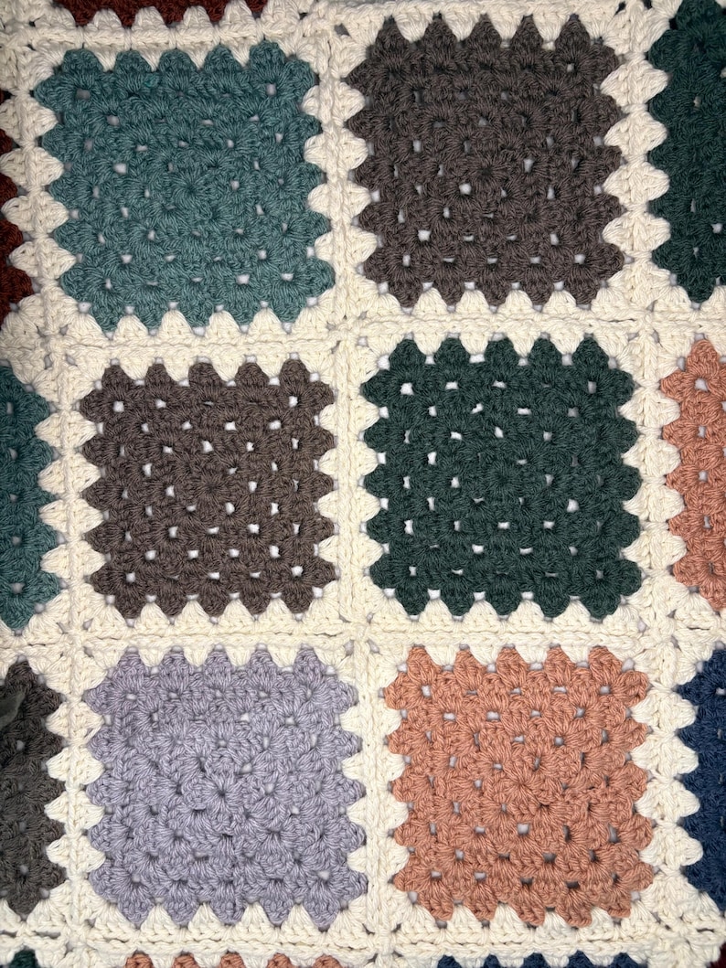 K&ouml;nnte beinhalten: Nahaufnahme einer handgefertigten H&auml;keldecke mit Patchwork-Design. Die Decke zeigt Quadrate in verschiedenen Farben, darunter T&uuml;rkis, Braun, Gr&uuml;n und Pfirsich, vor einem cremefarbenen Hintergrund. Die H&auml;kelstiche erzeugen ein strukturiertes, gem&uuml;tliches Aussehen.
