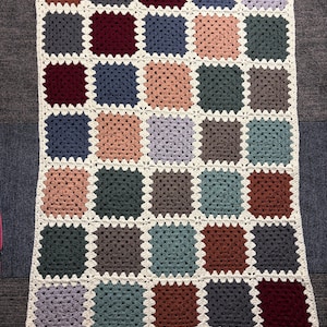 K&ouml;nnte beinhalten: Eine handgefertigte H&auml;keldecke mit Patchwork-Design. Die Decke besteht aus zahlreichen quadratischen Motiven in verschiedenen Farben, darunter Braun-, Blau-, Grau- und Rosat&ouml;ne, mit einem cremefarbenen Rand. Ungef&auml;hr 127 cm x 152 cm.