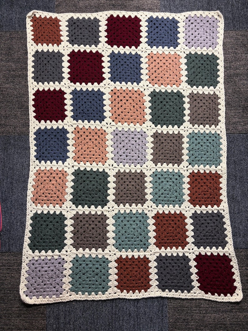 K&ouml;nnte beinhalten: Eine handgefertigte H&auml;keldecke mit Patchwork-Design. Die Decke besteht aus zahlreichen quadratischen Motiven in verschiedenen Farben, darunter Braun-, Blau-, Grau- und Rosat&ouml;ne, mit einem cremefarbenen Rand. Ungef&auml;hr 127 cm x 152 cm.