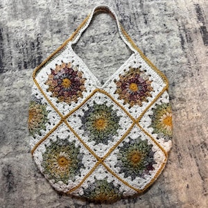 May include: A handmade crochet tote bag constructed from multiple square motifs featuring sunflower-like patterns in shades of olive green, mustard yellow, and deep plum, all bordered by white yarn and connected with a mustard yellow trim and handle.