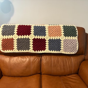 K&ouml;nnte beinhalten: Eine handgefertigte H&auml;keldecke mit einem Patchwork-Design aus bunten Quadraten. Die Quadrate sind in Graut&ouml;nen, Burgunderrot, Orange, Blau, Pfirsich und Gr&uuml;n gehalten, mit einem cremefarbenen Rand. Die Decke liegt auf einem braunen Ledersofa.