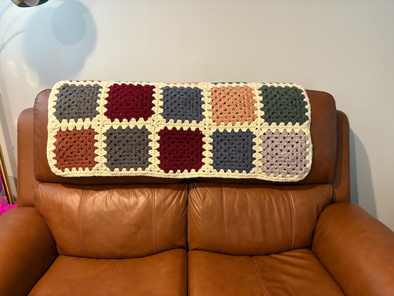 K&ouml;nnte beinhalten: Eine handgefertigte H&auml;keldecke mit einem Patchwork-Design aus bunten Quadraten. Die Quadrate sind in Graut&ouml;nen, Burgunderrot, Orange, Blau, Pfirsich und Gr&uuml;n gehalten, mit einem cremefarbenen Rand. Die Decke liegt auf einem braunen Ledersofa.