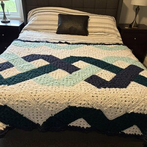 Puede incluir: Una manta de ganchillo hecha a mano en crema, verde azulado y azul marino, sobre una cama. La manta presenta un diseño geométrico con patrones entrelazados. La cama tiene un edredón a rayas, un cojín negro y dos mesitas de noche oscuras con lámparas.