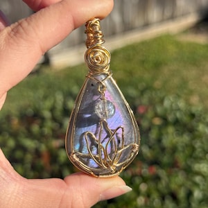 May include: A teardrop-shaped pendant with a gold wire wrap. The pendant features a shimmering, iridescent stone with purple and blue hues, and a small clear bead. A gold wire lotus flower design is visible.