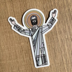 May include: A sticker featuring a stylized image of a religious figure with outstretched arms. The figure is depicted with a halo and dressed in light blue and gray robes. The design has a stained-glass effect, with black lines defining the shapes.
