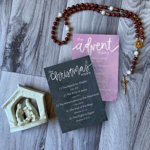 May include: A rosary with red and white beads, a gold crucifix, and a small nativity scene. Two cards read "The Advent Rosary" and "The Christmas Rosary" with scripture references. The items are arranged on a wooden surface.