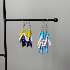 May include: Two pairs of dangle earrings made from small cylindrical beads. The first pair features yellow, white, purple, and black beads. The second pair features light blue, white, and light pink beads. Both pairs hang from silver-toned metal hooks.